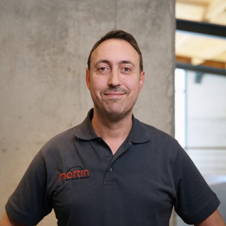 Ein Mann steht vor einer Betonwand, trägt ein graues Shirt mit Logo und lächelt freundlich.