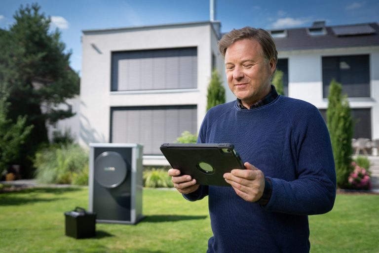 Mann mit Tablet steht im Garten neben einem Heizungsgerät und einem modernen Haus.